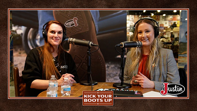 Two women talking to each other, sitting down behind a table, smiling at the camera.